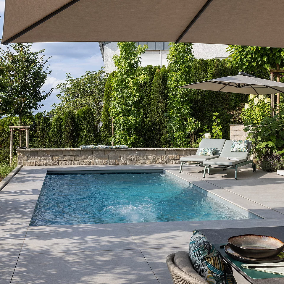 Moderner Garten mit Swimmingpool, Relaxliegen und großem Sonnenschirm neben einer Outdoor-Küche und gedecktem Tisch mit Geschirr und Obst.