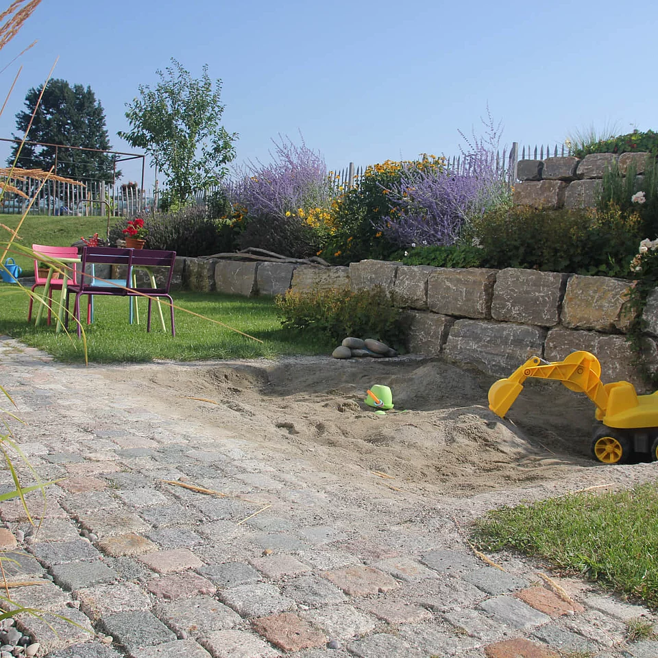 Sandkasten mit gelbem Spielbagger Sandkasten mit gelbem Spielbagger, Sandspielzeug und Grillplatz mit Tisch und Stühlen in einem Garten mit Steinmauer und Blumenbeet.