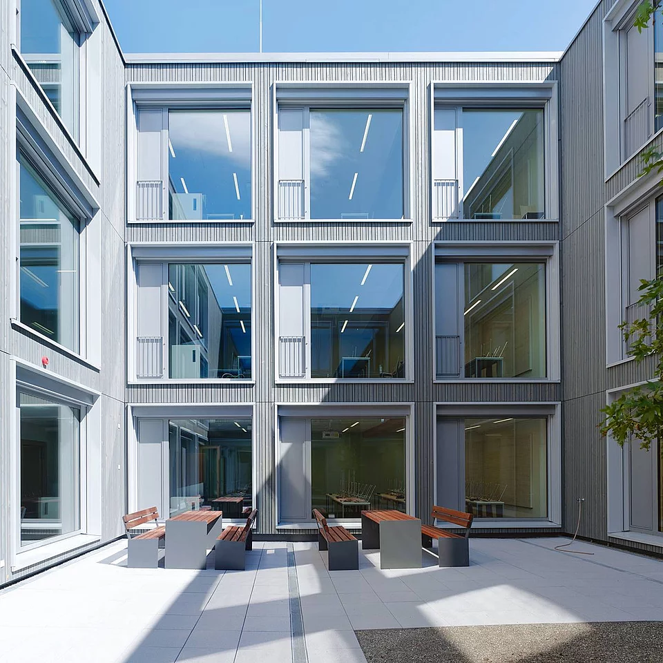 Polizeihochschule VS Moderner Innenhof mit großen Fensterfronten, Bänken und Tischen aus Holz und Metall, umgeben von einem mehrstöckigen Gebäude, mit einem Baum auf der rechten Seite.