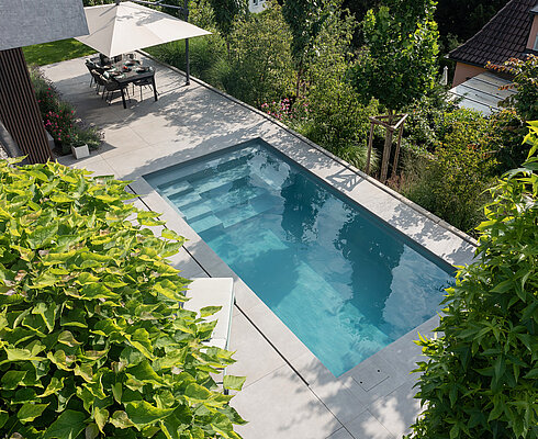 Moderner rechteckiger Swimmingpool in einem Garten, umgeben von grünen Pflanzen, mit einer Terrasse und einem Tisch mit Sonnenschirm im Hintergrund.