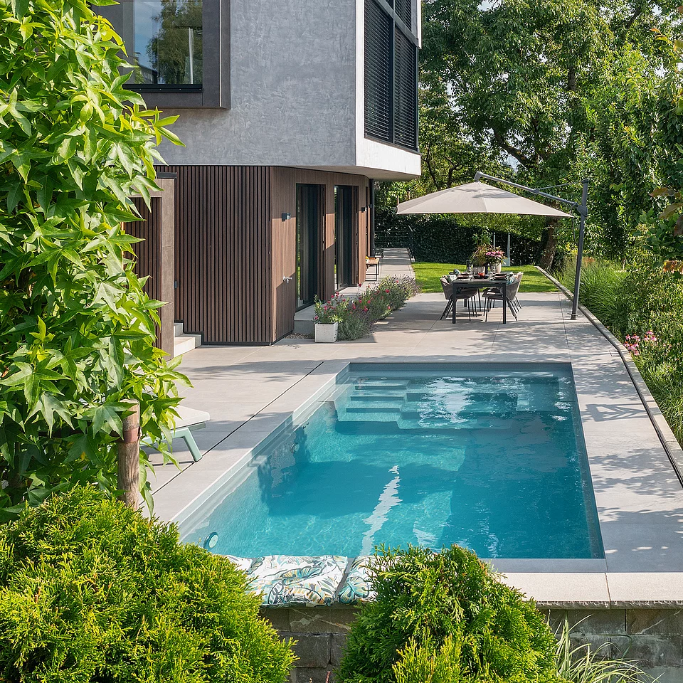 Moderner Garten mit kleinem rechteckigem Pool, angrenzender Terrasse mit Esstisch und Sonnenschirm, umgeben von grünen Pflanzen und Bäumen neben einem mehrstöckigen Haus.