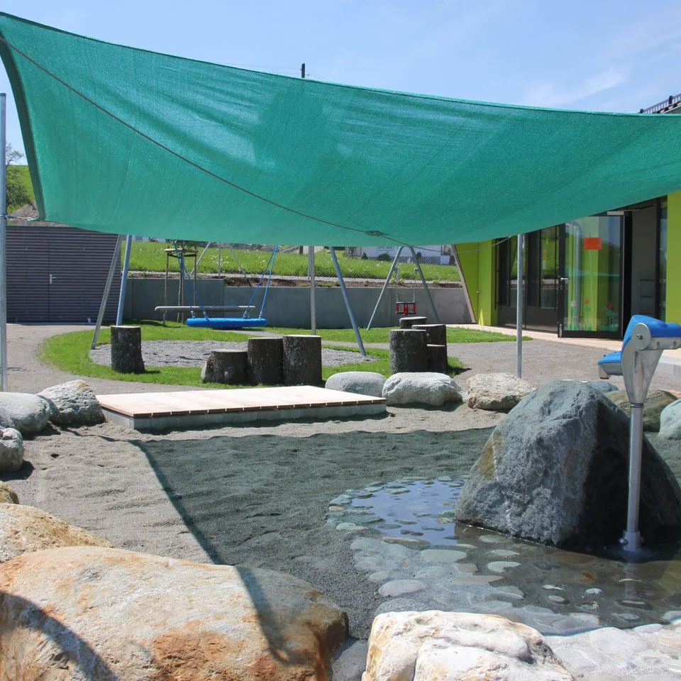 Freiraumgestaltung KIGA Rötenbach Außenbereich mit Sandkasten und Wasserpumpe, darüber grünes Sonnensegel gespannt, im Hintergrund Spielplatz mit Schaukeln und Sitzhockern aus Baumstämmen.