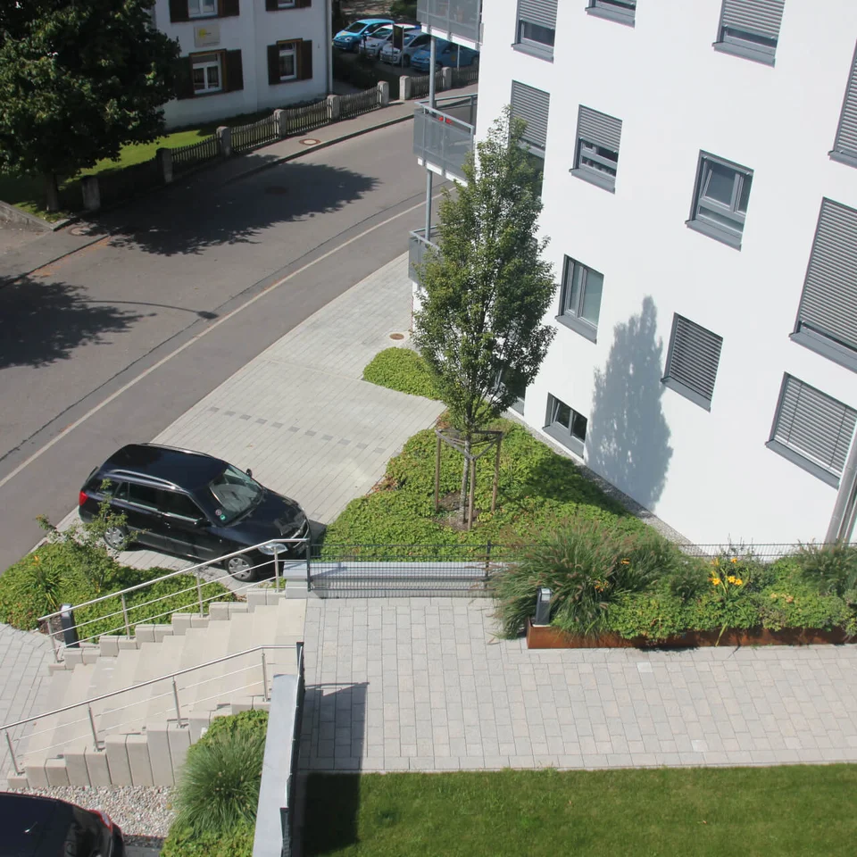 Freiraumgestaltung Wohnumfeld Tettnang Moderne Wohnanlage mit weißem Gebäude, Gehweg, Grünflächen und zwei parkenden Autos in sonniger Umgebung.
