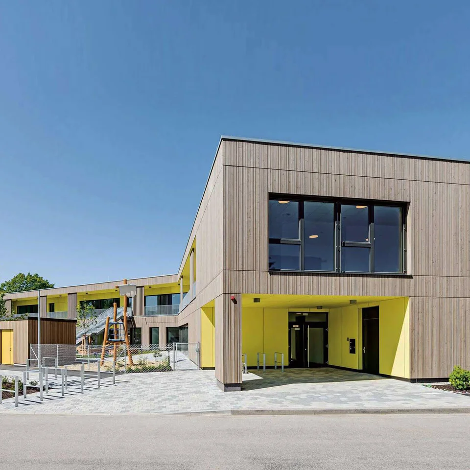 Gestalteter Außenbereich Kita Schäferhof Modernes, helles Kita-Gebäude mit Holzfassade und großem Eingangsbereich bei sonnigem Wetter.
